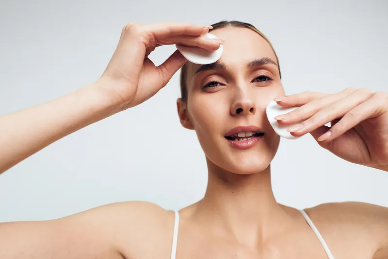 A woman uses cotton pads to remove makeup, performing a gentle facial cleanse in a bright studio. Closeup portrait illustrating skincare, cleansing, and self care.