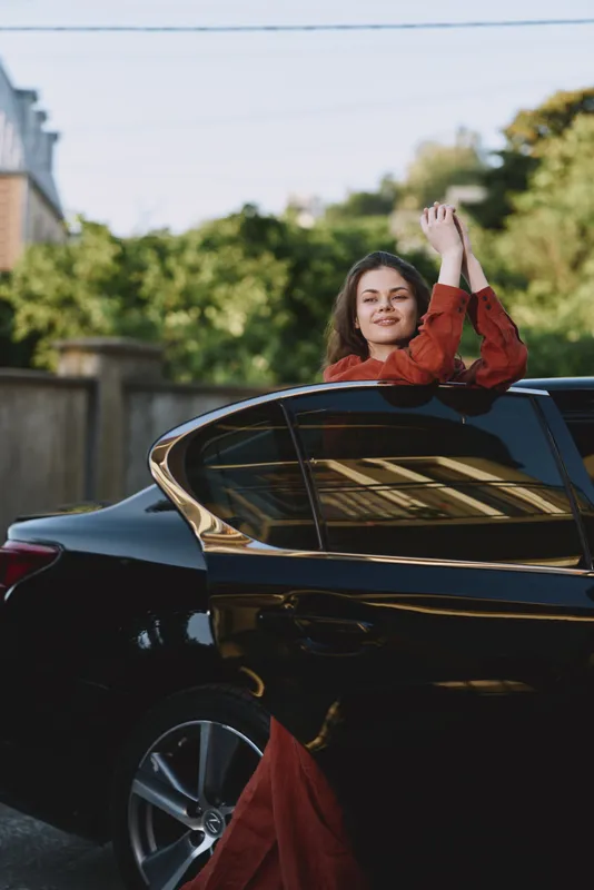 A woman in a red dress leans from a sleek car, waving with a joyful smile in daylight. A candid lifestyle moment blending fashion, motion, and urban energy
