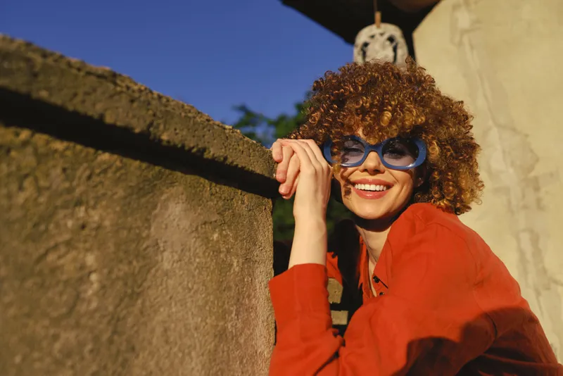 Smiling woman with curly hair in a red top leans on a sunlit wall outdoors, wearing stylish sunglasses. A bright, candid lifestyle moment capturing confidence and warmth