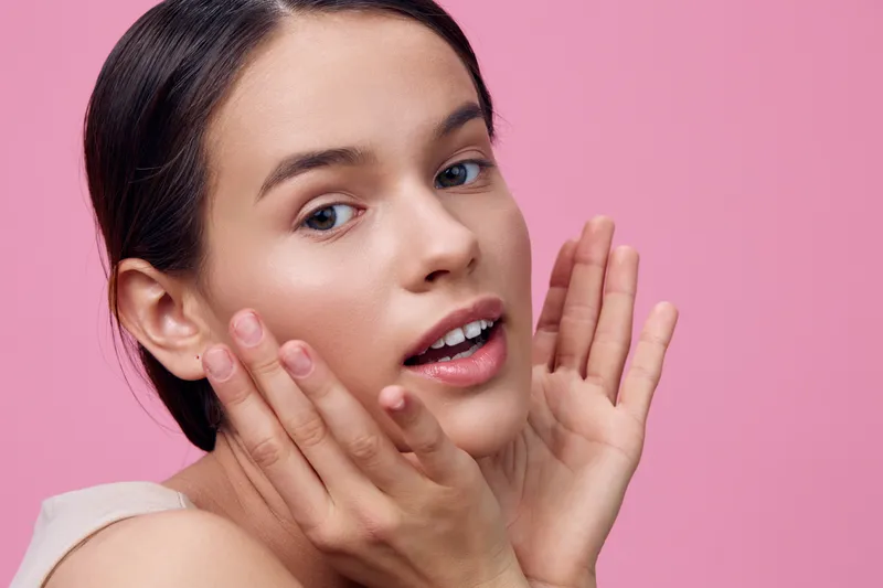Young woman with clear skin, expressing joy and beauty, posing against a soft pink background, ideal for health and beauty concepts