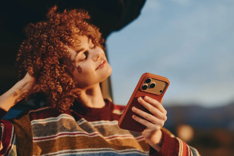 Lifestyle portrait of a woman with curly hair holding a smartphone outdoors, touching her cheek as she takes a selfie, communication and technology concept in warm evening light casual sunset