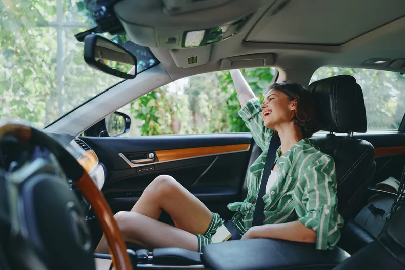 A joyful passenger sits inside a car, smiling under daylight, wearing casual attire as the vehicle interior and seatbelt frame a bright, relaxed moment.
