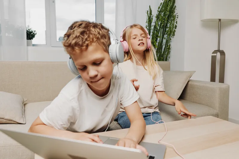 Two children with headphones enjoy music and technology at home, boy focused on laptop and girl feeling relaxed with eyes closed on sofa.