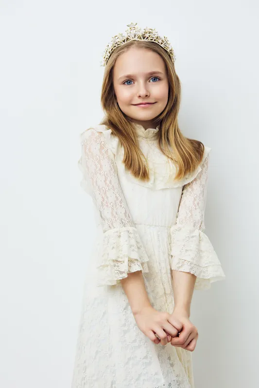 Joyful young girl in a cream lace dress with a crown, exuding innocence and charm in a minimalistic studio setting Ideal for children s fashion or portraits