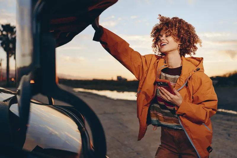 Woman lifestyle on a beach at sunset, wearing an orange jacket, standing by a car door as a person reaches out, travel mood, curly hair visible, portrait feeling