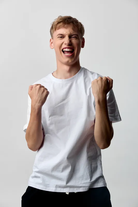 Energetic man in a white tee celebrates triumph with clenched fists and a wide smile, conveying victory, motivation, and high spirits in a casual studio setting