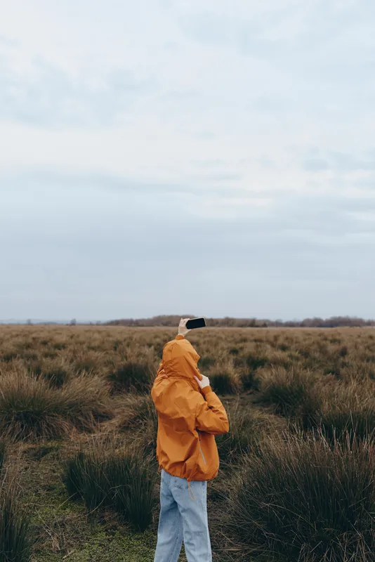 Woman lifestyle outdoors field grass jacket backpack, standing in a vast meadow, looking toward cloudy sky, calm mood, natural light, casual travel outfit, slow walking concept alone
