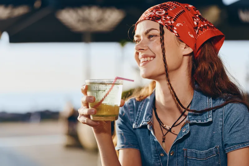 Lifestyle woman in modern boho-western denim street style smiling and enjoying a refreshing drink with warm film color tones and natural outdoor lighting.