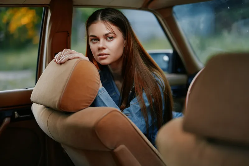 Young woman with long hair sitting in a vintage car, wearing a denim jacket, with a contemplative expression, capturing a nostalgic and relaxed mood.