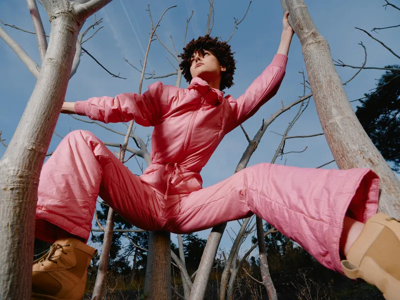 Outdoor fashion jumpsuit worn by a model mid jump among trees, showcasing a dynamic pose and youthful energy in a natural setting with bright sky and bare branches