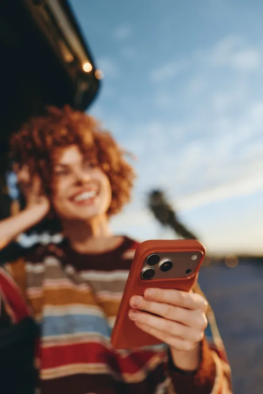 Woman lifestyle portrait outdoors on beach in summer, curly hair woman smiles while holding smartphone red case, selfie near ocean waves, travel mood, bright sunlight, soft blur