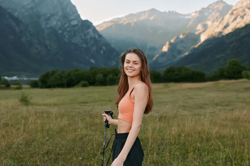 A cheerful woman stands outdoors in a scenic valley with mountains in the background. She wears an orange sports top and dark pants, smiling at the camera while holding a rope, enjoying fresh air and