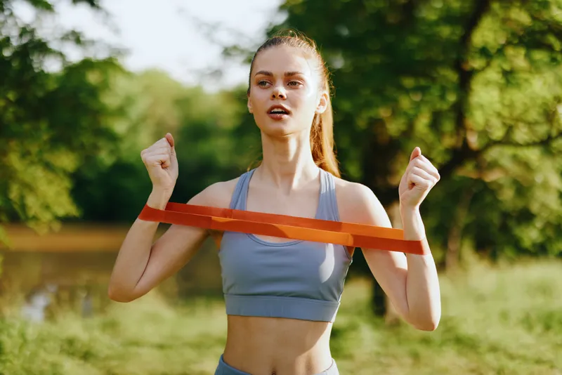 Young woman exercising outdoors with resistance bands, showcasing determination and fitness in a natural green background, perfect for healthy lifestyle themes