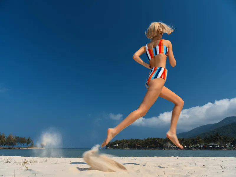 A woman in a colorful striped bikini leaps mid air on a sunny beach, sand spraying behind her, turquoise sea in the distance and a clear blue sky above.