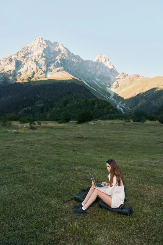 A young woman sits on a grassy meadow with a laptop, set against rugged mountain scenery and a clear blue sky. She looks focused, calm, and productive outdoors.