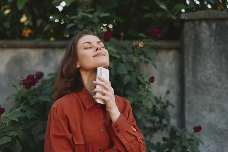 Young woman in a rust shirt enjoys a serene outdoor moment, eyes closed, leaning against a garden wall while holding a smartphone near her cheek amid blooming roses