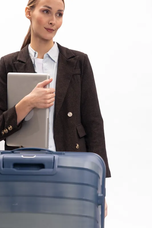 Young businesswoman in blazer holding laptop stands near blue suitcase on white background. Concept of business travel, professionalism, and modern work lifestyle with confident expression.