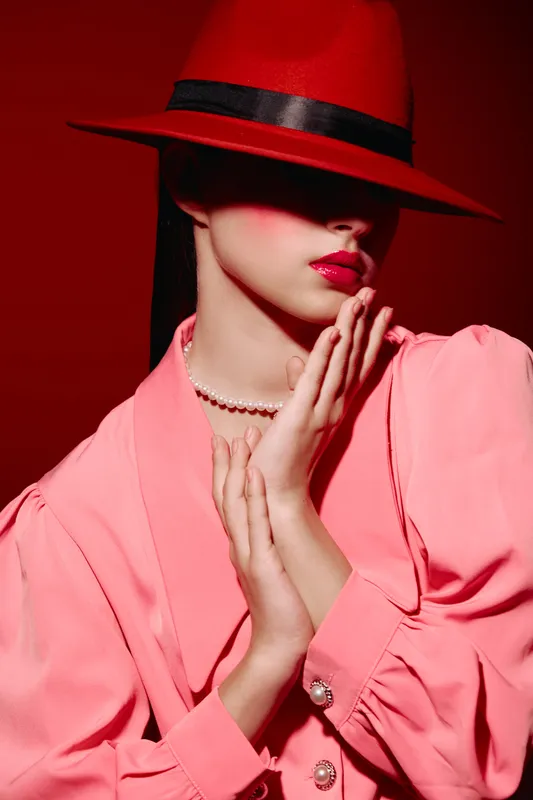 Artistic portrait of a fashion model wearing a red hat and pink blouse with pearl buttons. Soft lighting highlights red lipstick and elegant hands near the face. Beauty concept.