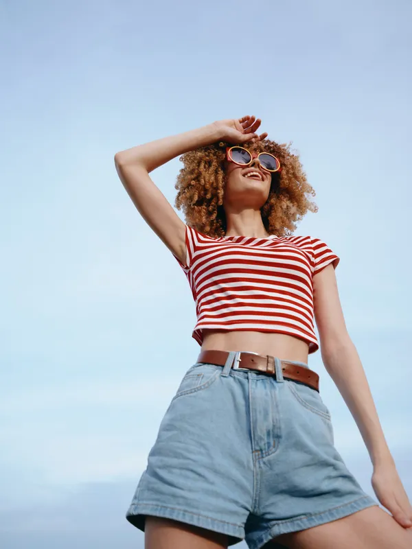 A stylish young woman with curly hair wearing a red and white striped crop top, denim shorts, and sunglasses stands against a bright blue sky, exuding carefree summer vibe.