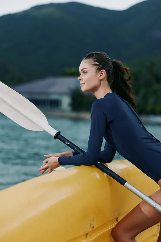 Young woman in a blue wetsuit paddles a yellow kayak along a calm lake, with distant mountains and lush scenery, conveying balance, motion, and outdoor adventure.