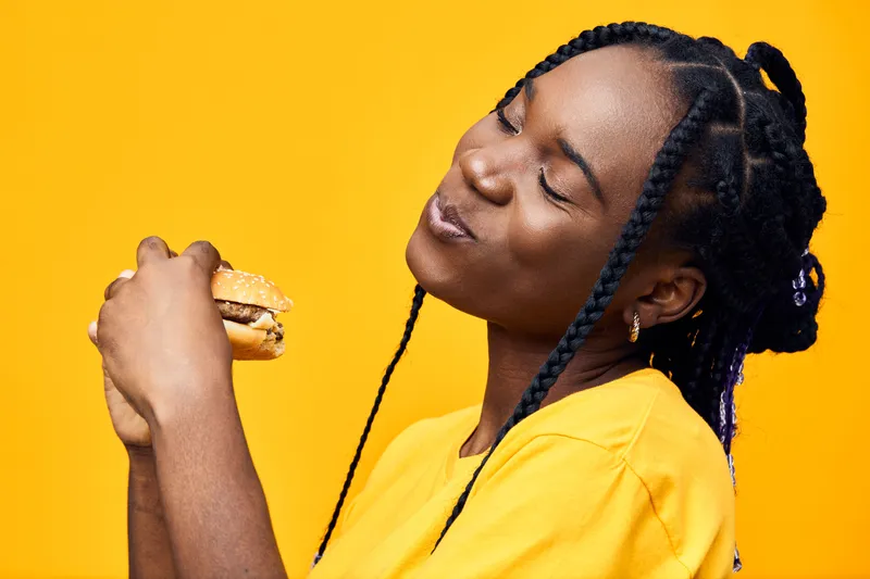 Joyful young woman enjoying a delicious burger against a vibrant yellow background, highlighting happiness and indulgence in healthy eating habits