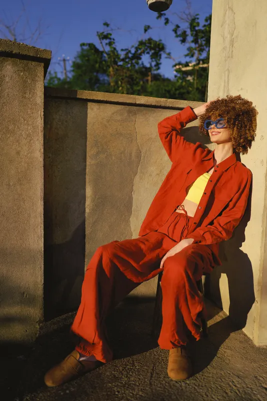 Cheerful woman outdoors in bold red and orange attire, relaxed pose under bright sun, sunglasses, sunlit wall, warm tones, natural greens in background, carefree lifestyle moment