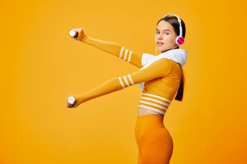 Woman in bright orange sportwear lifts dumbbells with headphones against a vivid orange backdrop, showcasing fitness, exercise, movement and energetic sport-inspired training.