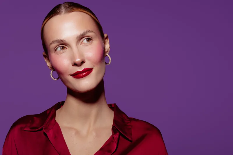 Young woman with red lipstick and hoop earrings looks sideways on purple background. She wears a red blouse and has a confident, thoughtful expression. Beauty and fashion concept.