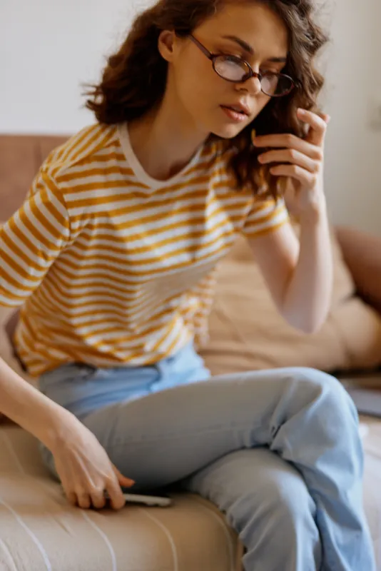 Young woman in a striped shirt sits on a cozy home sofa, casually scrolling on her phone with a thoughtful, relaxed mood under soft natural light in a simple everyday scene.
