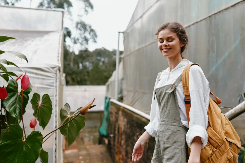 Smiling young woman in stylish overalls exploring a greenhouse, surrounded by vibrant plants, showcasing a connection with nature and sustainable living