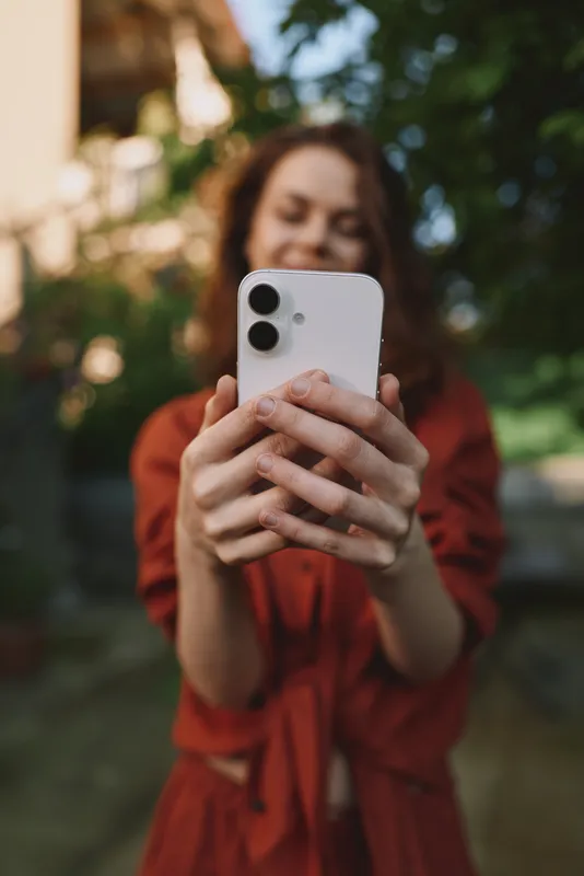 Smiling woman takes a casual selfie outdoors with a smartphone, capturing a relaxed lifestyle moment in natural light against a garden backdrop, candid and vivid