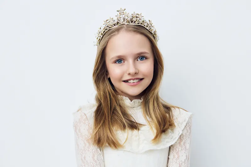 Charming young girl with a crown, wearing a vintage white dress, smiling sweetly, against a light gray background, perfect for themes of childhood and innocence
