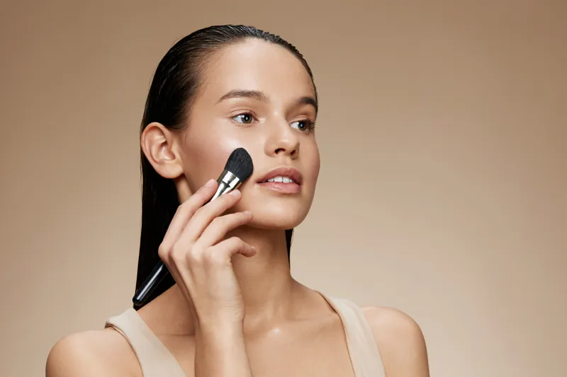 Beautiful young woman applying makeup with a brush on a neutral background, highlighting natural beauty, skincare, and self care practices