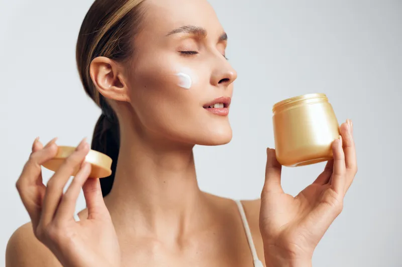Woman applying cream from a gold jar, close up portrait, skincare routine in progress, soft lighting, dewy complexion, calm expression, attentive moment, indoors, clean background, natural glow,