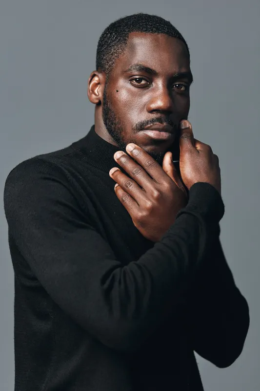 African American man with dark skin wearing black turtleneck sweater posing with hands near face in creative visual designer style studio portrait with gray background and serious expression.