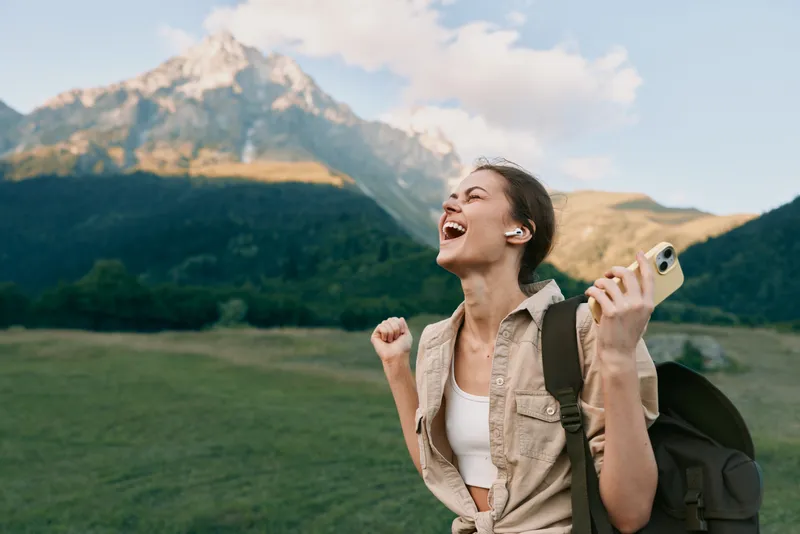 Joyful woman outdoors, hiking in mountains, backpack on shoulder, smartphone in hand, smiling with sunlit sky, open meadow, lifestyle moment, natural colors.