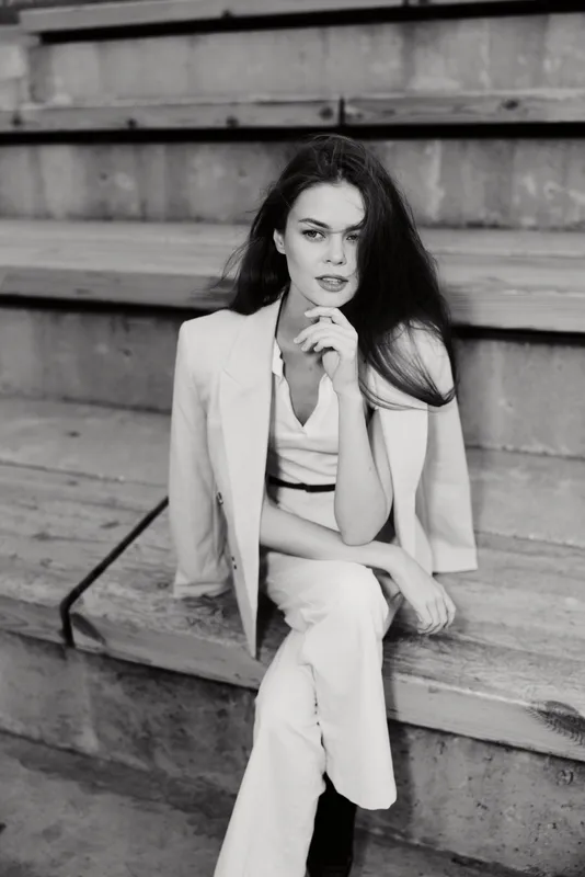 Woman in white business suit sitting on stairs, thoughtful expression, black and white fashion portrait with stylish elegance and confidence