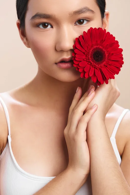 asian woman holds a red gerbera close to her face in a soft studio portrait, calm expression, natural lighting highlighting smooth skin and subtle makeup.