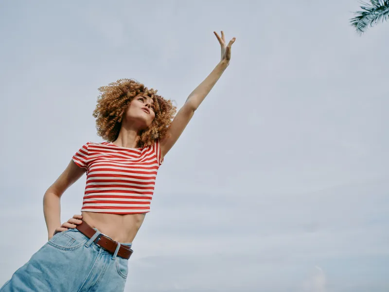 A carefree young woman with curly hair wears a red and white striped crop top and high waisted denim jeans, dancing against a clear blue sky.