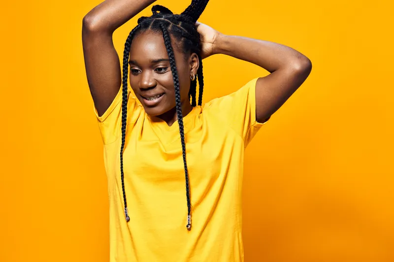 Young Black woman with braided hair smiling while adjusting her hair against a vibrant yellow background, showcasing a lively and energetic mood