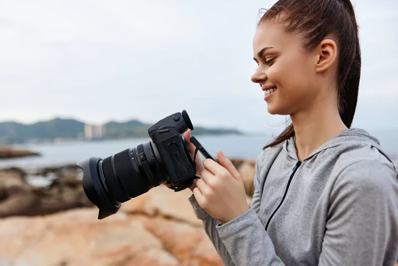 Young woman with a camera smiling, dressed in a gray hoodie, enjoying outdoor photography by the sea, capturing the beauty of nature