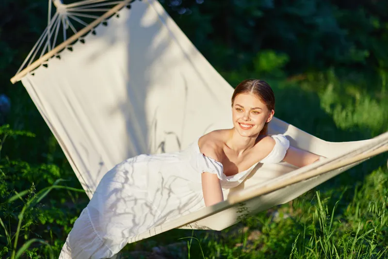 Young woman relaxing in a hammock outdoors, wearing a flowing white dress, smiling in a natural green setting, evoking a sense of tranquility and summer joy