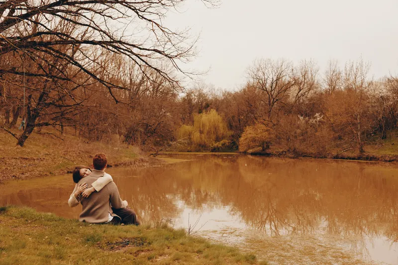 Couple outdoors by a calm lake in autumn, sharing a quiet moment among colorful trees. Warm, soft light and tranquil water create a serene mood of connection, reflection, and gentle affection in a