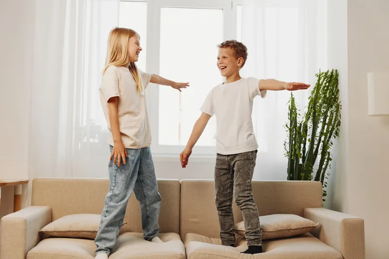 Two happy children standing on a sofa in a bright living room, smiling and reaching out to each other. Concept of playful siblings, joy, and family bonding at home.