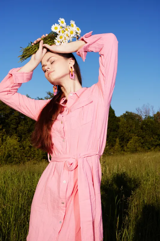 Young woman in a pink dress holding daisies, enjoying nature in a vibrant summer field, expressing joy and tranquility against a blue sky