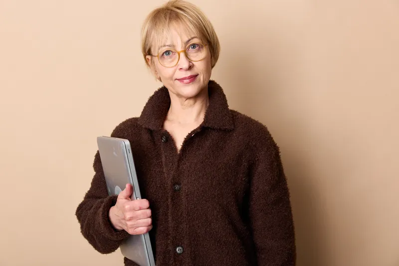 Elderly woman with glasses in studio portrait holding documents against beige background calm expression concept of office work support and communication