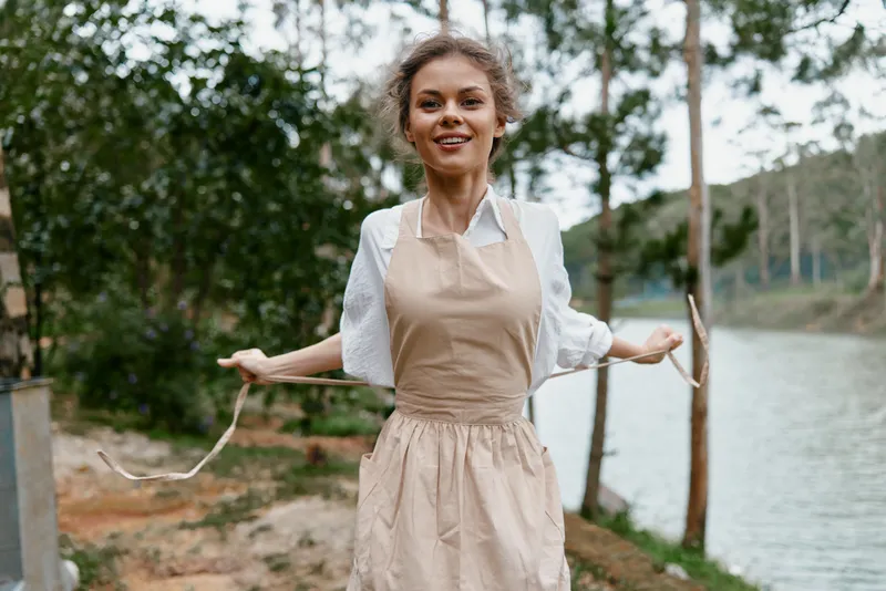 Young woman in a beige apron joyfully skipping near a serene lake, surrounded by greenery, showcasing happiness and a love for nature during the outdoor spring season