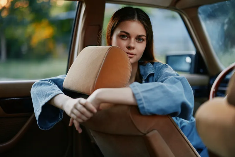 Young woman in a vintage car, wearing denim jacket, looking thoughtfully out of the window, evoking a sense of nostalgia and freedom during sunset.