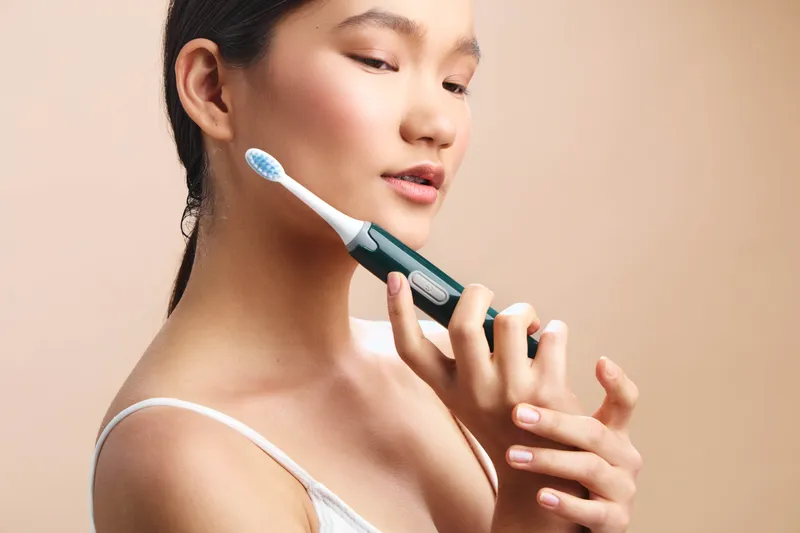 Asian woman smiles as she uses an electric toothbrush, practicing daily dental care in a bright bathroom, highlighting healthy habits and confident self care in a modern lifestyle.