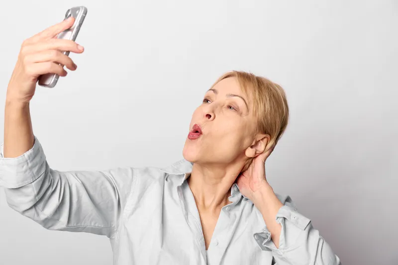 Woman holding a smartphone for selfie, touching ear and neck, portrait with calm wellness and beauty, on bright white background, soft studio light, closeup